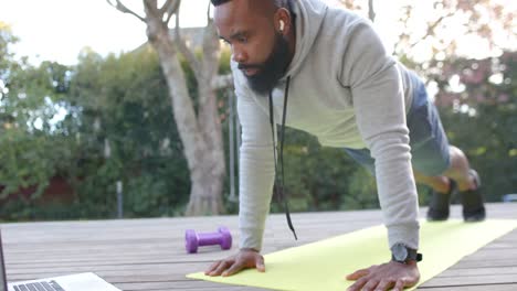 Hombre-Afroamericano-Enfocado-En-Entrenamiento-Físico-Haciendo-Flexiones-En-Cubierta-En-Un-Jardín-Soleado,-Cámara-Lenta