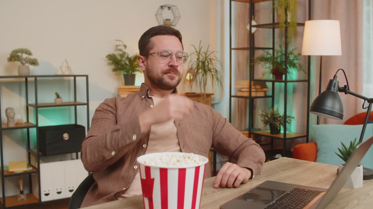 joven empresario feliz tomando un descanso del trabajo comiendo palomitas de maíz y viendo películas en la computadora portátil en casa