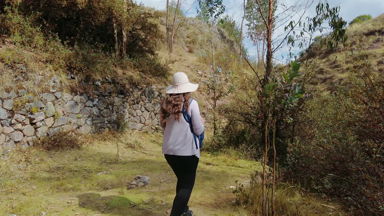 A woman treks along an ancient Inca road in the mountains near Cusco, passing stone ruins and dense vegetation. The trail is part of the historic Qhapaq Ñan network.