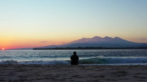 Silhouette-of-girl-sitting-on-exotic-beach-in-front-of-big-sea-waves-watching-sunset-with-magical-sky-colored-on-yellow,-purple-and-red-in-Indonesia