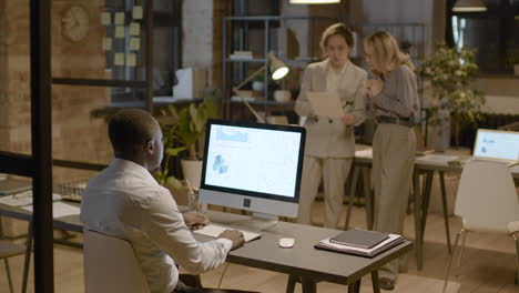 Rear-View-Of-American-Man-Worker-Sitting-At-Desk-While-Watching-Stadistics-And-Graphics-On-Computer-Monitor