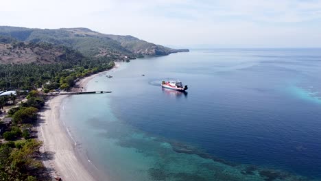 Rising-aerial-drone-of-passenger-ferry-leaving-Atauro-Island-tropical-island-over-stunning-coral-reef-ecosystem,-turquoise-ocean-waters-and-rugged-coastal-landscape,-Timor-Leste,-South-East-Asia