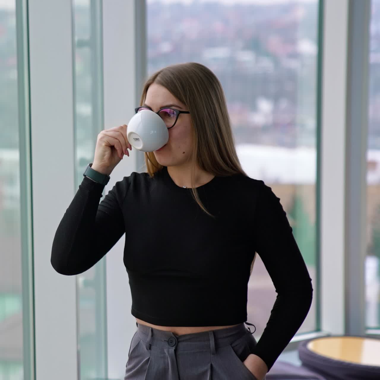 Business lady with a cup of coffee indoors. Young woman in glasses having a break while standing near the window in office and drinking hot beverage