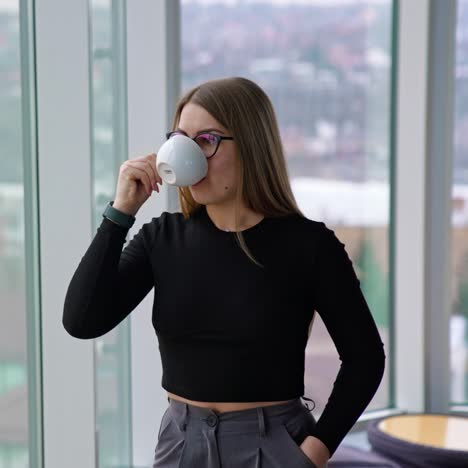 business lady with a cup of coffee indoors. young woman in glasses having a break while standing near the window in office and drinking hot beverage