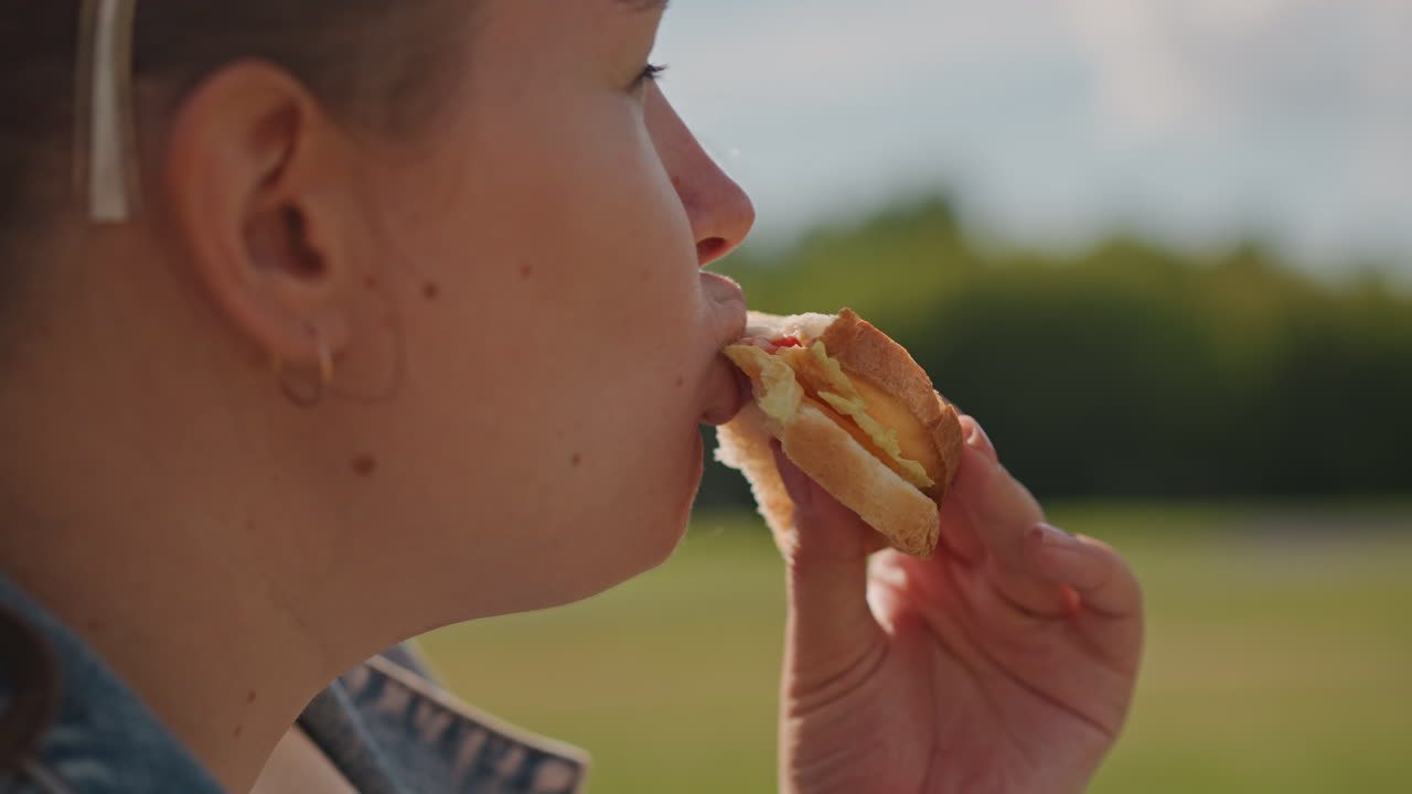 White woman biting sandwich in profile, closeup of hand and bread with soft field background, earrings and headband visible, slow contemplative bite, texture and crust focus, warm summer light, quiet