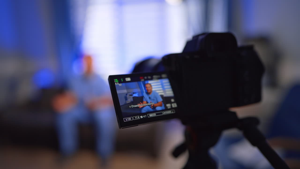 Man in blue uniform talking at the little display of the modern camera. Doctor recording a vlog in his office. Selective focus