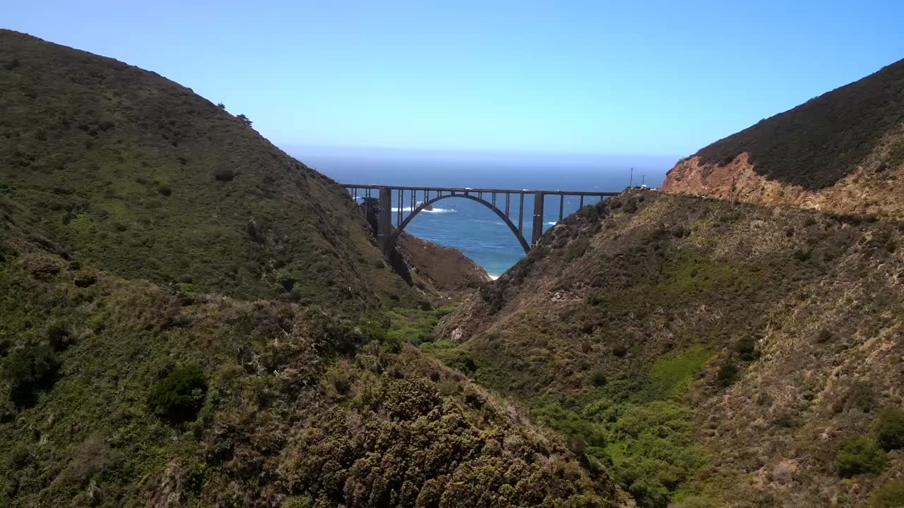 빅 서 (big sur) 에 대한 영화적인 공중 드론 촬영, 두 산 정상을 연결하는 다리를  ⁇ 처하고 배경에는 넓은 파란 바다가 있습니다.