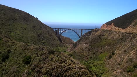 Cinematic-aerial-drone-shots-over-Big-Sur,-capturing-the-bridge-linking-two-mountain-peaks-with-the-expansive-blue-ocean-in-the-background