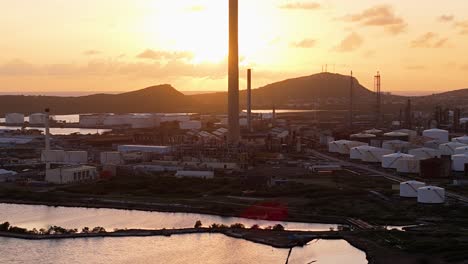 golden hour glow in sky lights up behind flare stacks and large storage tanks of oil refinery