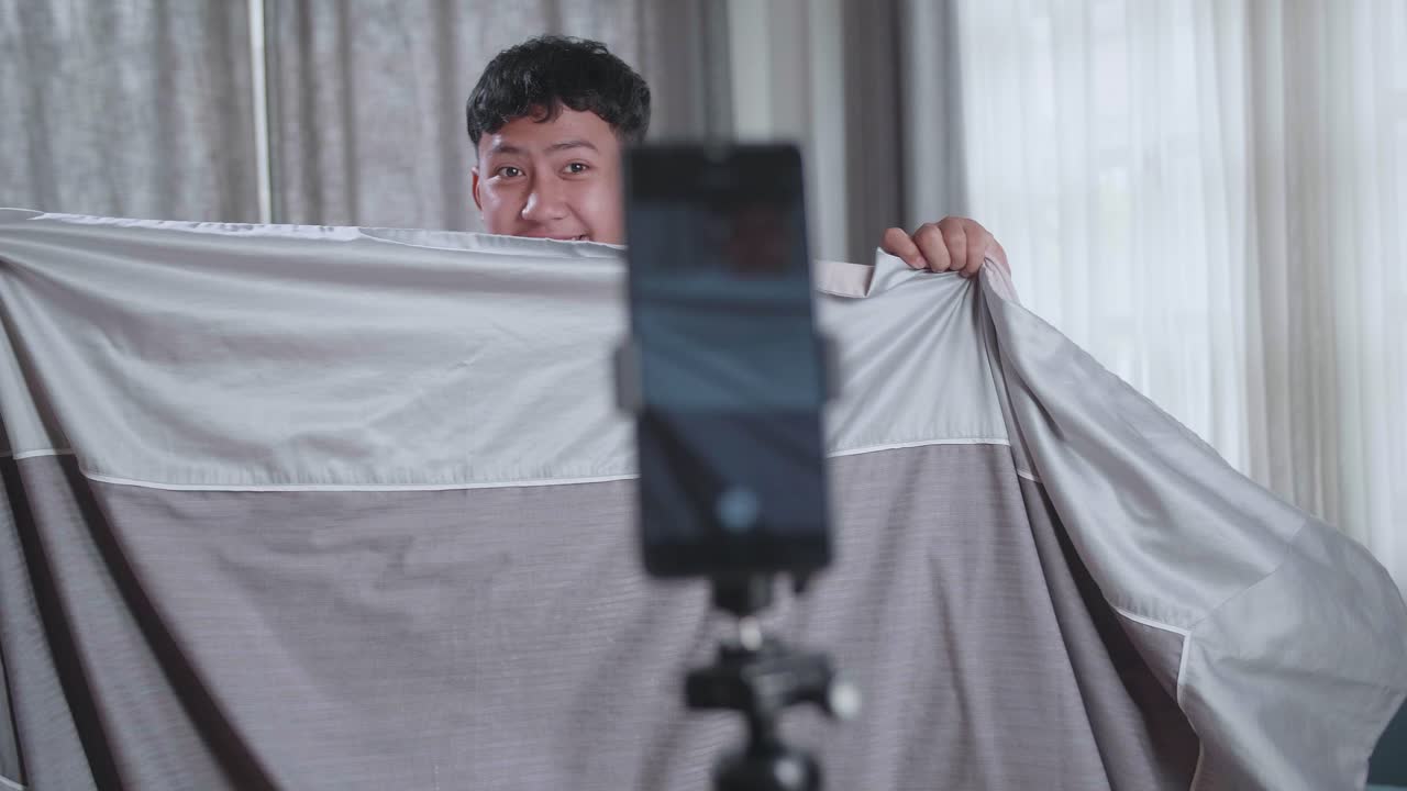 Display Smartphone Of Asian Boy Shows Trick With Disappearing On Mobile Phone Camera While Recording Video Content For Social Networks In Cozy Room