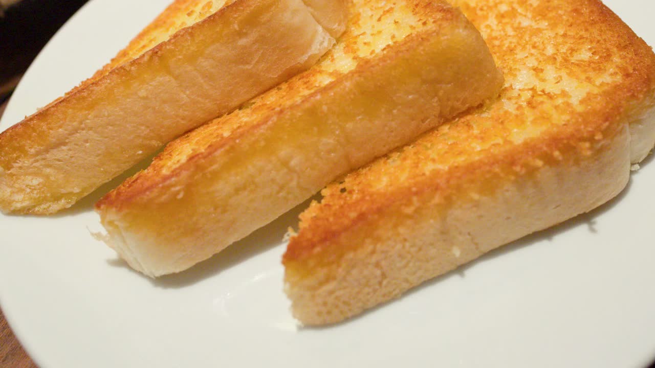 Close-up of hand picking toasted garlic bread slices from white plate under warm lighting