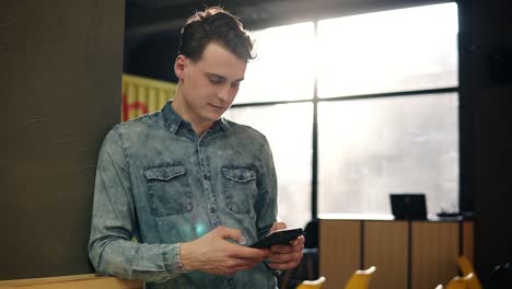 Young-Attractive-Sexy-Guy-In-Denim-Shirt-Standing-And-Scrolling-Something-On-His-Smartphone