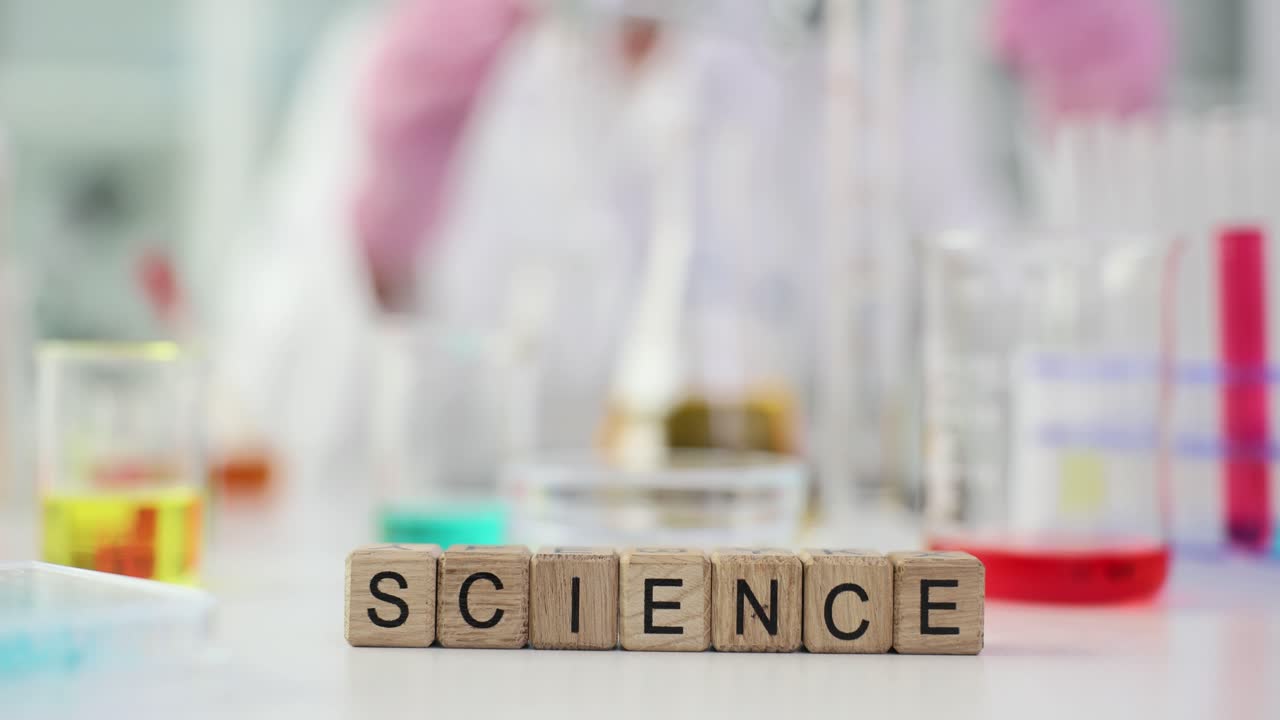 Wooden blocks spelling 'SCIENCE' in a laboratory setting with beakers and a blurry scientist