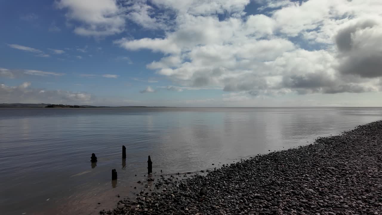 Litorale della baia di Morecambe con ciottoli scuri, acqua calma che riflette le nuvole e vecchi pali di legno.
