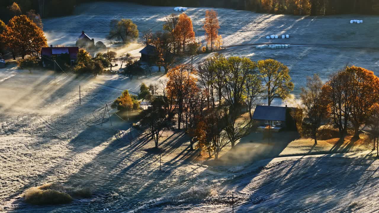 Frosty rural landscape with farmhouses and autumn trees, illuminated by morning sun rays