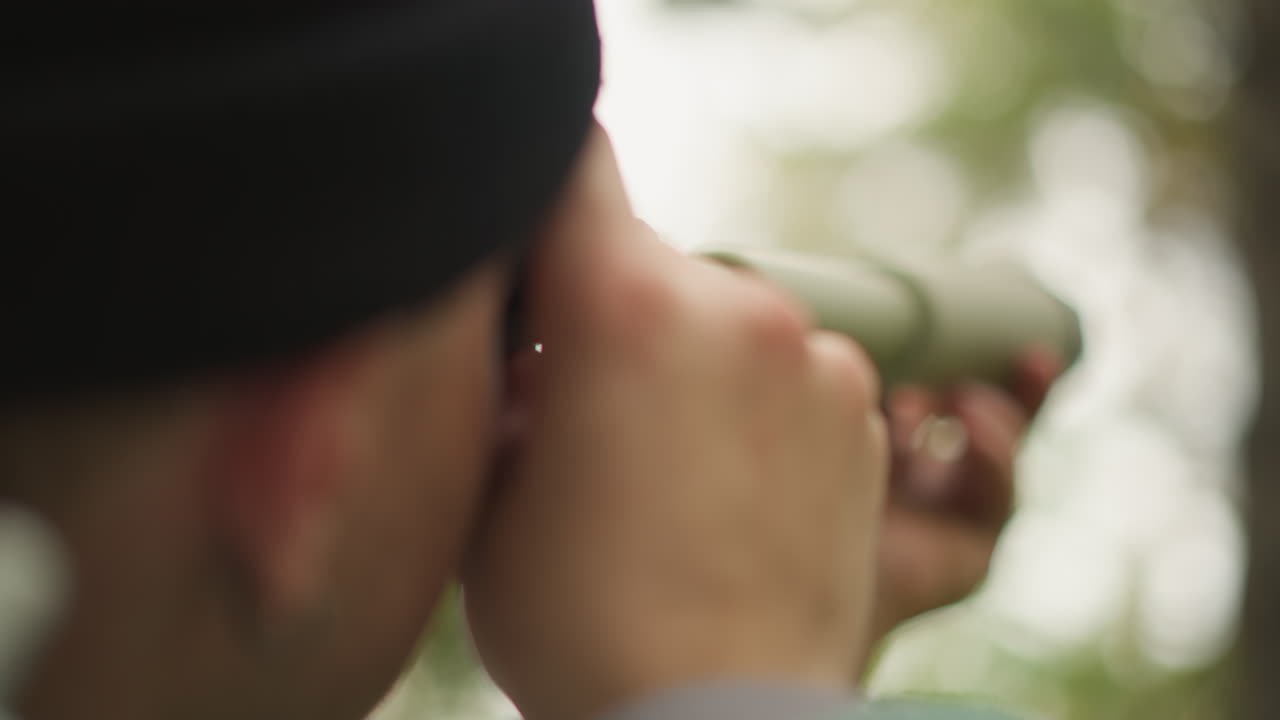 traveler peers through monocular into forest, close profile and beanie visible with steady hand grip on tube, focused observation and quiet scouting action, scout or birdwatcher role in green