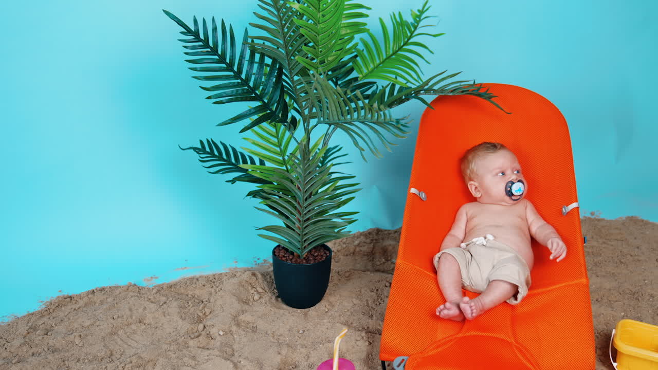 Sweet little baby in shorts and with pacifier lies on the orange chair. Beach sand and plant pot around the kid.