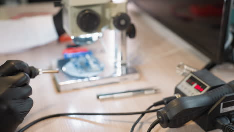 a close-up view of a hand wearing a black glove operating a soldering station in a lab, the setup includes a microscope with a circuit under it and various electronic tools on a wooden desk