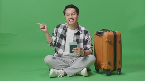 full body of asian male traveler with luggage and passport smiling and pointing to side while sitting in the green screen background studio