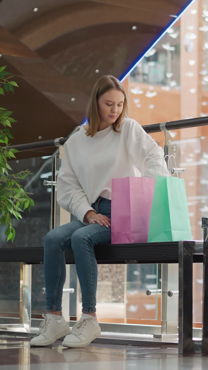 joven sentada en un centro comercial moderno y brillante recupera un libro cubierto de amarillo de una bolsa de compras rosada, rodeada de bolsas de colores, plantas decorativas, barandillas de vidrio, con reflejos de luz dispersos