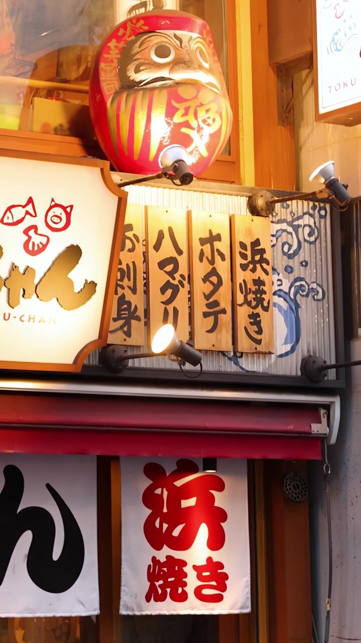 La telecamera si inclina verso l'alto mostrando la facciata di un tradizionale izakaya di Tokyo con menu esposti e una bambola Daruma