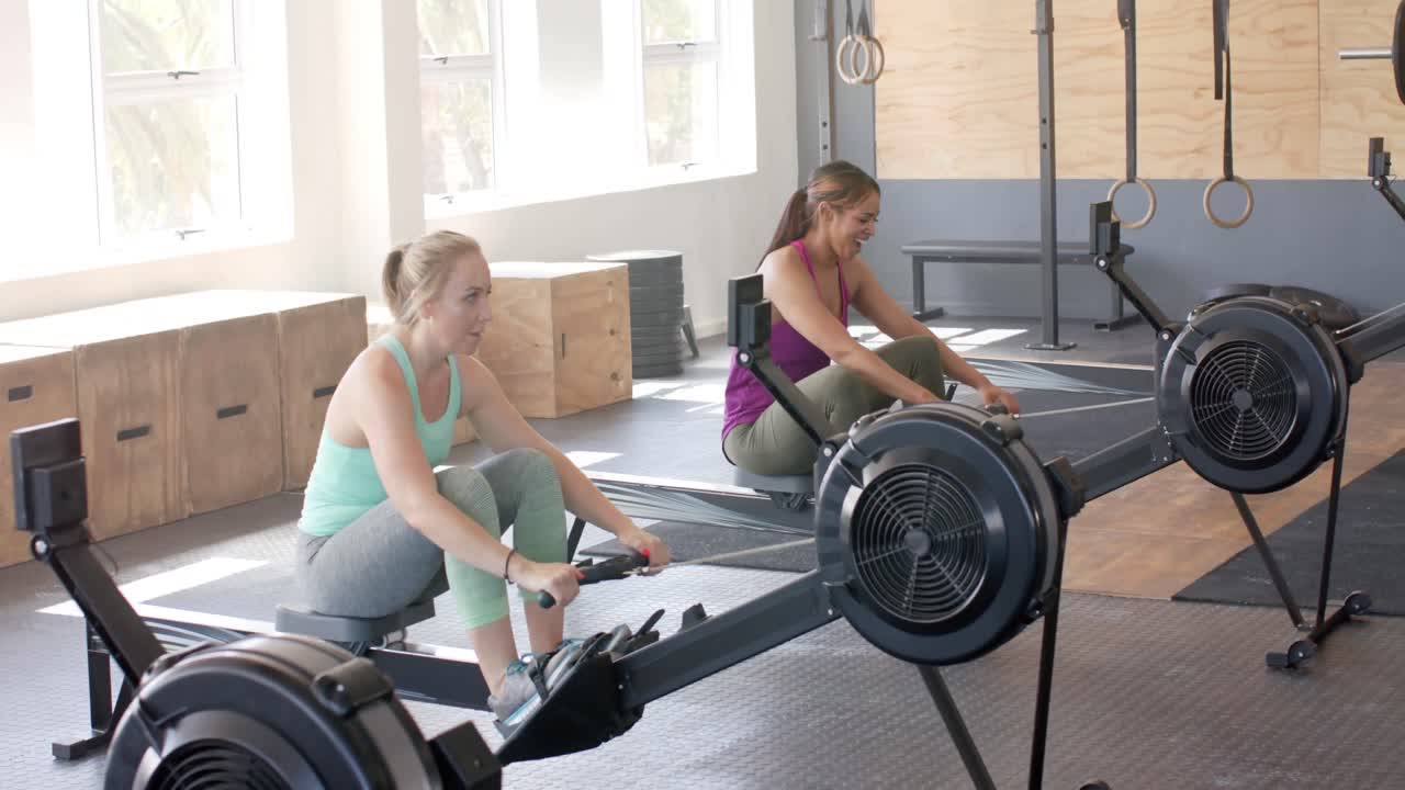 Happy unaltered diverse women fist bumping after exercising on rowing machines at gym, slow motion