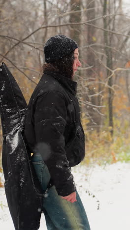 joven con chaqueta de invierno con las manos en los bolsillos y la guitarra en la espalda caminando por una ruta cubierta de nieve en la nevada, rodeada de árboles desnudos y nieve cayendo