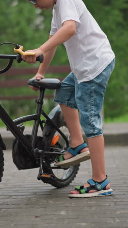 se ve a un niño con una camiseta blanca y pantalones cortos de vaqueros ajustando el soporte de su bicicleta negra, tratando de estacionarla en un camino pavimentado en un parque