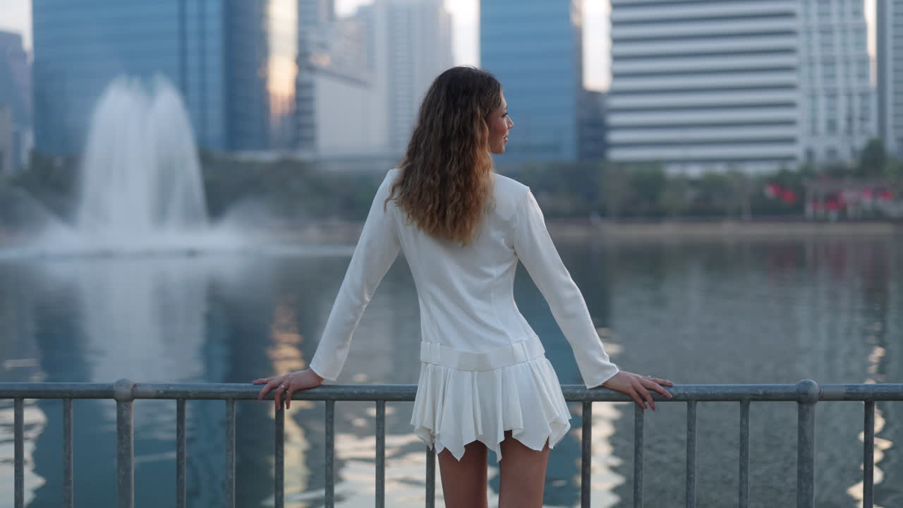 Woman in white dress looking at city lake with fountain and buildings
