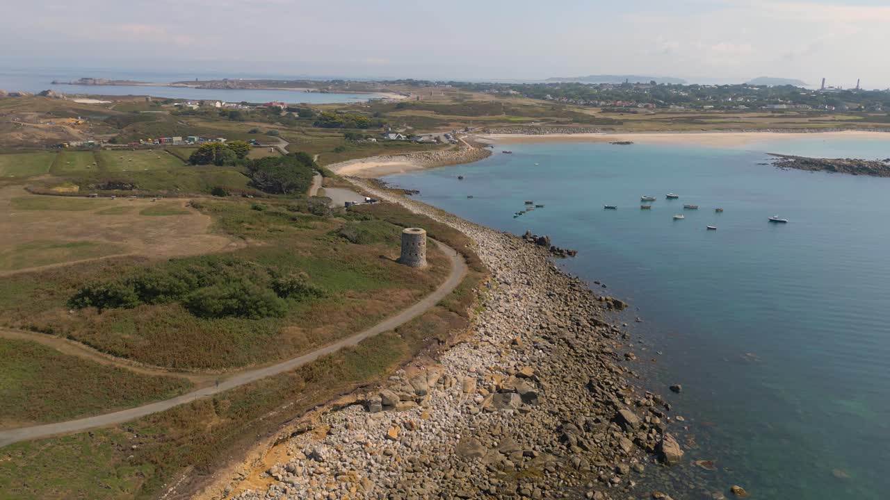 Circling flight centered around historic Martello Tower on coastal promontory with golden beaches,rocky foreshore and boats at anchor in a clear turquoise sea