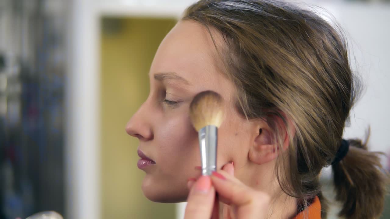 Side view of a young, pretty female model. Make up process. Artist putting some transparent, shiny rouge on woman's cheek using