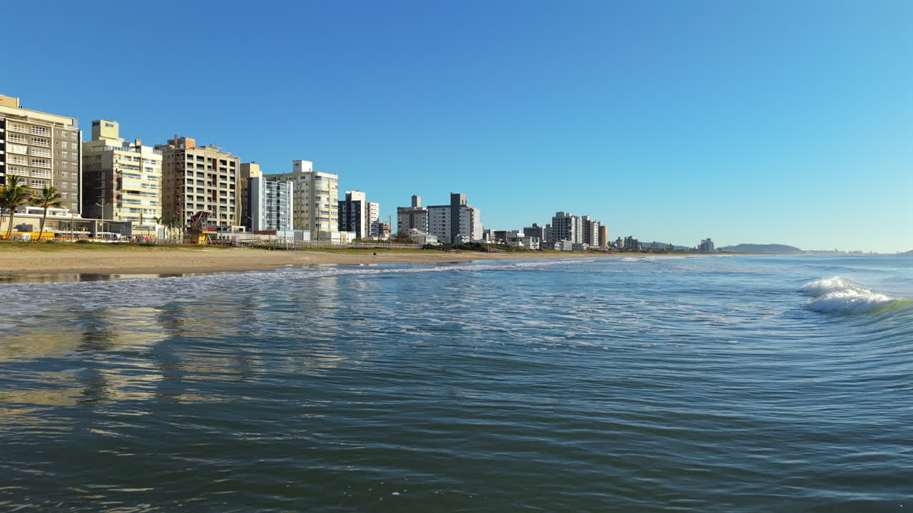 Drone shot of ocean waves reaching the sandy shore of Navegantes, Brazil, with seaside buildings in view
