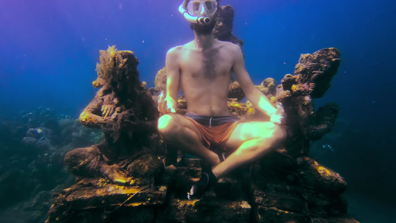Giovane uomo con maschera e boccaglio che medita sotto il mare accanto ad alcune statue asiatiche sommerse