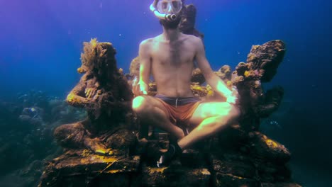 joven con máscara y snorkel meditando bajo el mar junto a algunas estatuas asiáticas sumergidas