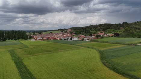 an aerial drone view ,flying backward, of a small spanish village inside soria province, surroended by green wheat fileds, under a dramatic stormy skies.