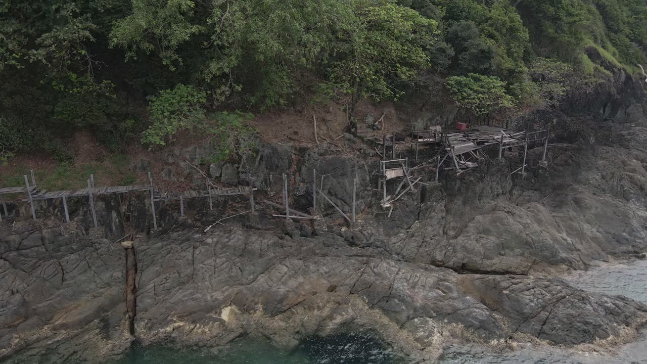 Drone trucking orbit shot , birds eye view of a abandoned derelict wooden board walk and deck along granite rock coastline on a tropical Island with jungle lush green forest and ocean
