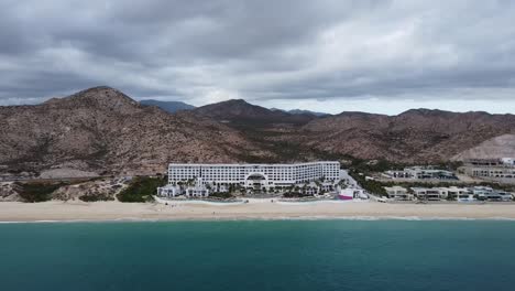 Luftschwenkaufnahme-Vor-Dem-Luxushotel-Des-Marquis-Los-Cabos-Hotels-Mit-Blick-Auf-Die-Küstenlandschaft-Mit-Traumhaftem-Strand,-Türkisfarbenem-Meerwasser-Und-Majestätischen-Bergen-Im-Hintergrund