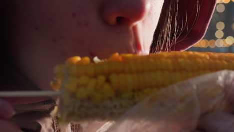 close-up of girl eating corn held by stick wrapped in white plastic with bokeh lights in blurred background, steam rising from fresh corn in outdoor setting, enjoying street food snack