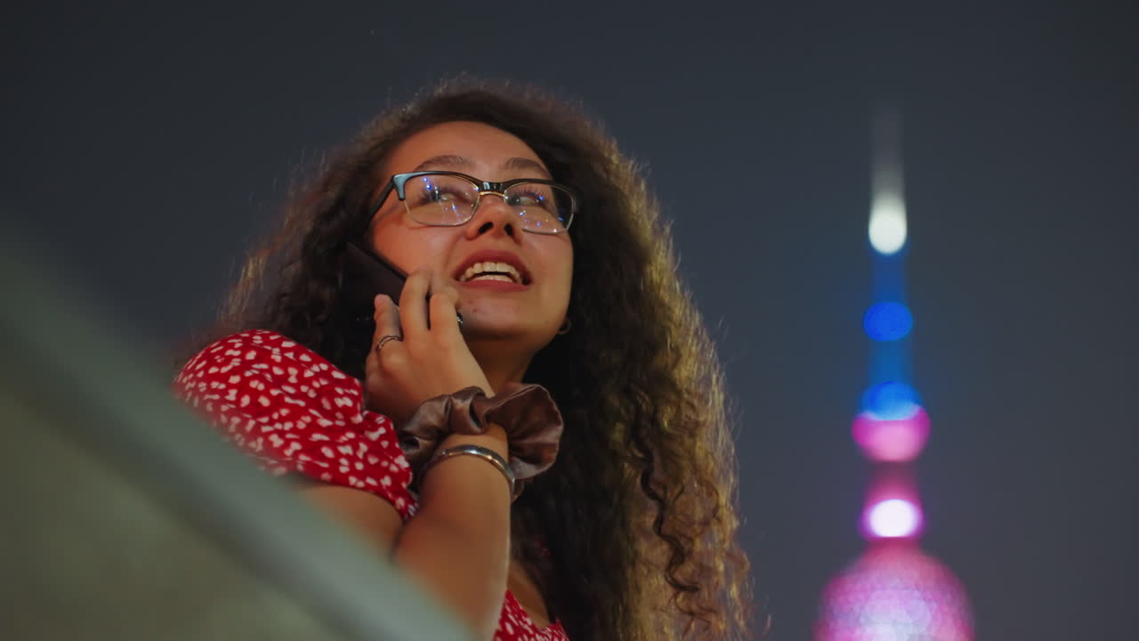 Femme souriante parlant au téléphone sur un balcon en ville, bokeh de tour néon en arrière-plan, cheveux bouclés et lunettes, robe rouge à motifs, bracelets, conversation joyeuse et spontanée, téléphone portable tenu à la main, lueur chaleureuse d'un lampadaire.