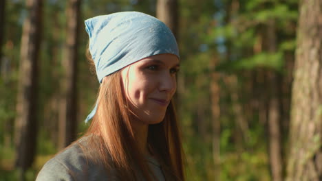 close-up of woman with blue headscarf standing in sunlit forest surrounded by tall trees, a gentle smile reveals her dimples as sunlight reflects softly off her face