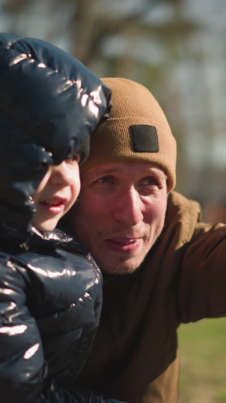 아버지가 그의 아들을 다리 위에 들고 앉아있는 모습, 아버지가 미소 짓고 멀리 있는 무언가를 흥분하게 가리키고, 아버지가 헤드 워머를 착용하고 있습니다.