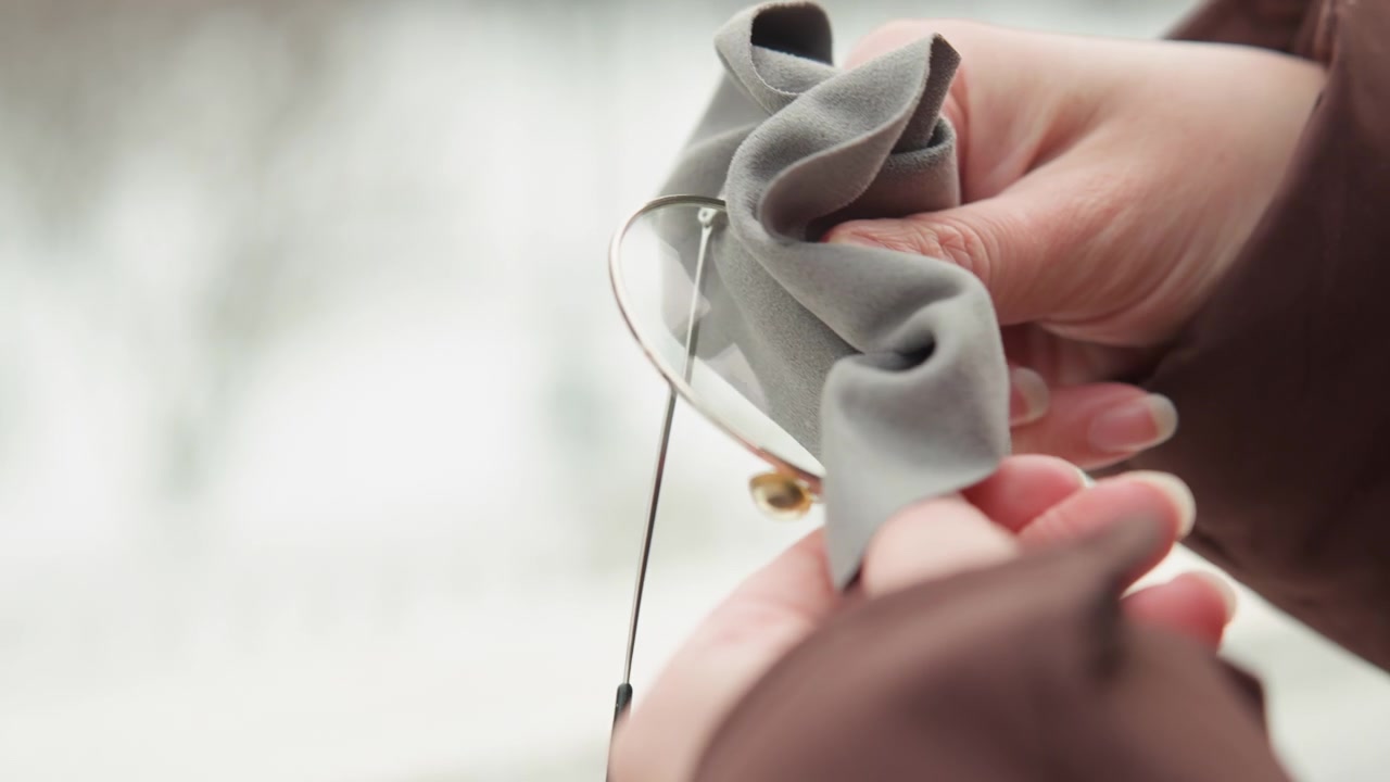 Hands gently lift thin metal rim eyeglasses from textured soft fabric case, fingertips holding frame while case folds inward, with delicate chain, soft daylight, tactile detail, shallow blur, quiet