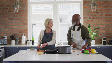 Una-Pareja-De-Ancianos-De-Raza-Mixta-Usando-Delantales-Cocinando-Comida-Juntos-En-La-Cocina-De-Casa