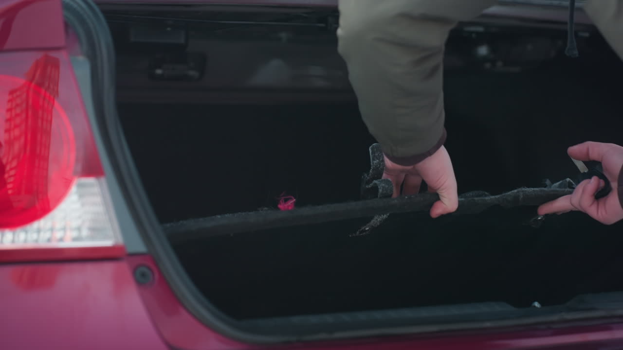 Rear view of person in green winter jacket retrieving spanner from car boot and placing it on black interior cover, during cold day outdoors, trunk open and backlight