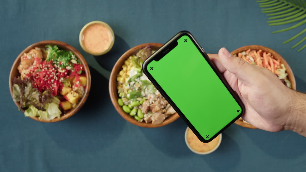 Hawaiian poke bowls, holding smartphone with chroma green screen top view. Cooked poke made of sliced vegetables, seafood and greenery. Healthy vegetarian dishes. Asian vegan raw meal, chopsticks