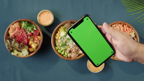 hawaiian poke bowls, holding smartphone with chroma green screen top view. cooked poke made of sliced vegetables, seafood and greenery. healthy vegetarian dishes. asian vegan raw meal, chopsticks