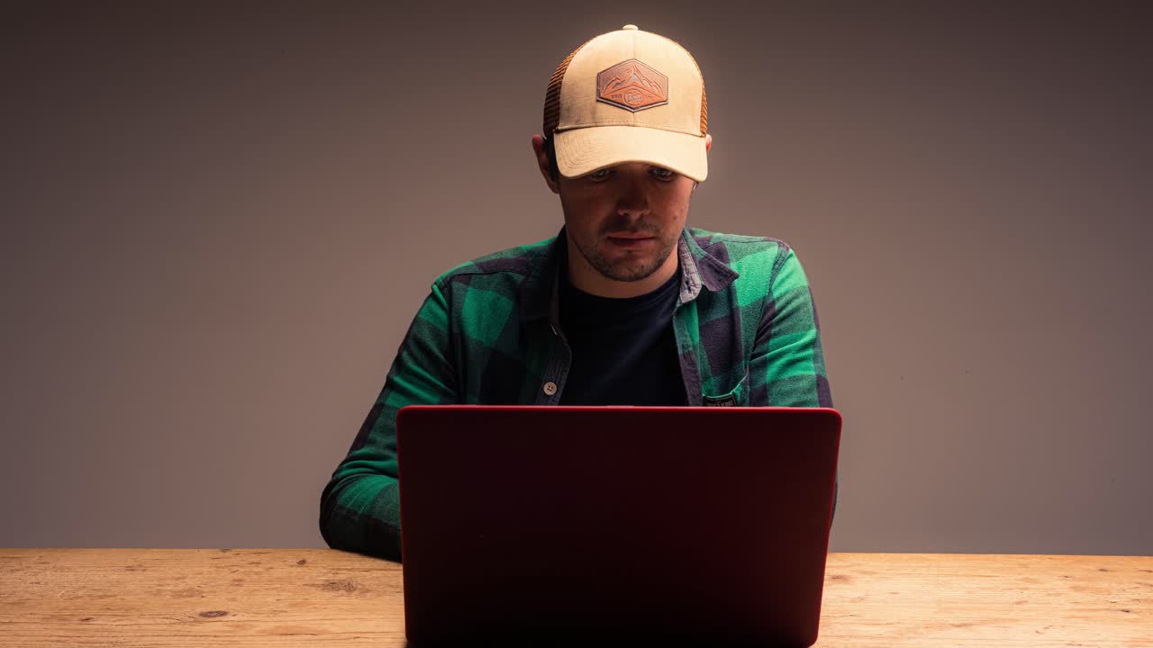 Junger Mann mit Hut arbeitet am Computer auf grauem Hintergrund.
