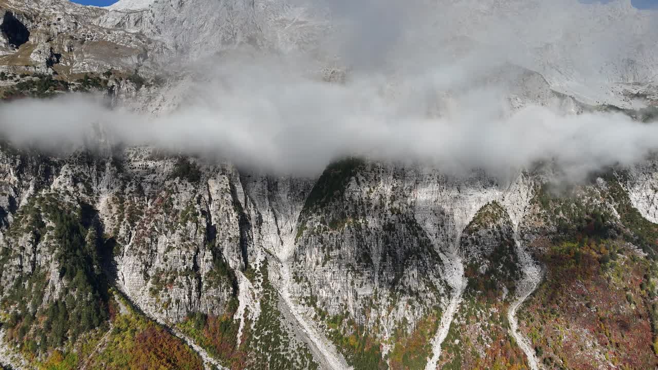 Drone shot of Veliki Movran mountain with rugged cliffs partly covered by drifting clouds. Sunlight highlights rocky textures and tree-covered lower slopes in remote wilderness area
