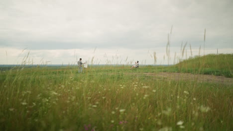 an artist, wearing a hat and checkered shirt, is focused on painting in a serene meadow filled with tall grass under a cloudy sky. in front of him, a woman sits peacefully in a chair