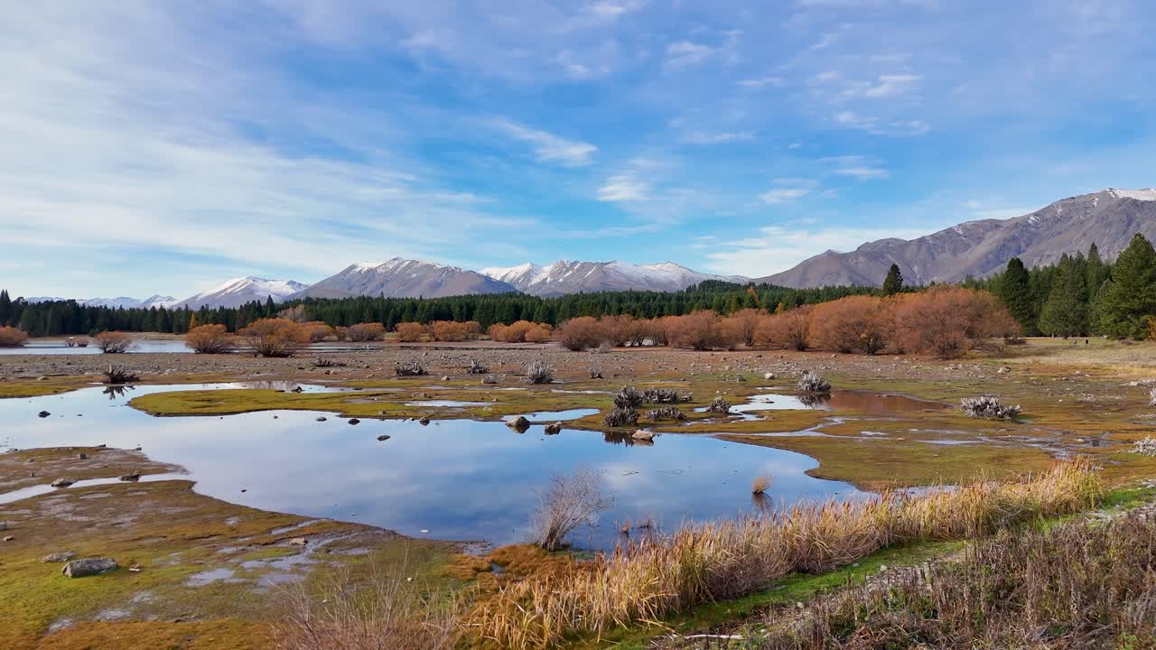 Drone footage captures Lake Tekapo's reflective waters and autumn landscape with mountains in the background under a clear sky